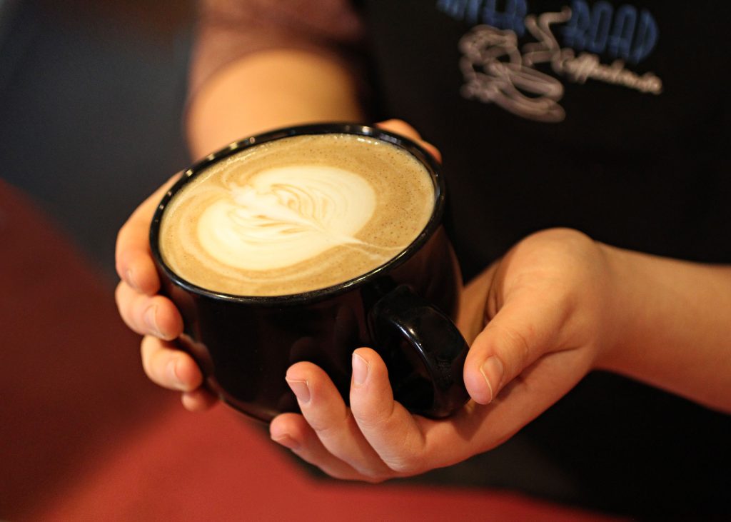 An up-close photo of a person holding a black coffee mug with both hands. The coffee is a latte with a foam design.