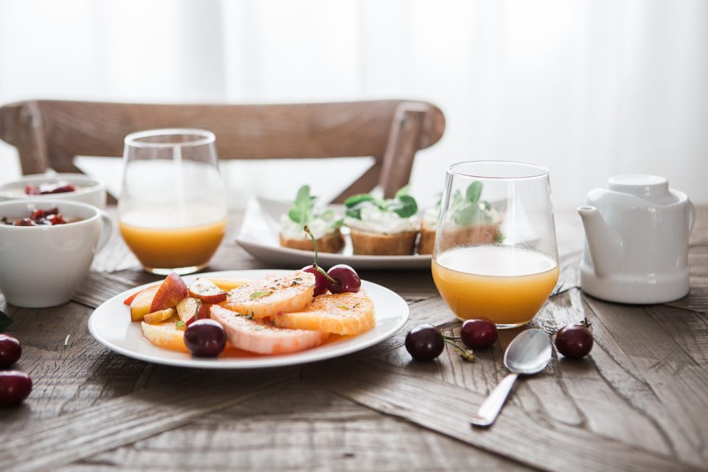 Two drinking glasses with orange juice and a breakfast spread of fruit.