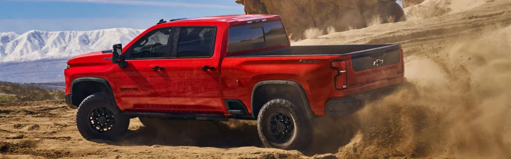 A red Chevrolet Silverado ZR2 driving through sandy terrain with snowy mountains in the background.