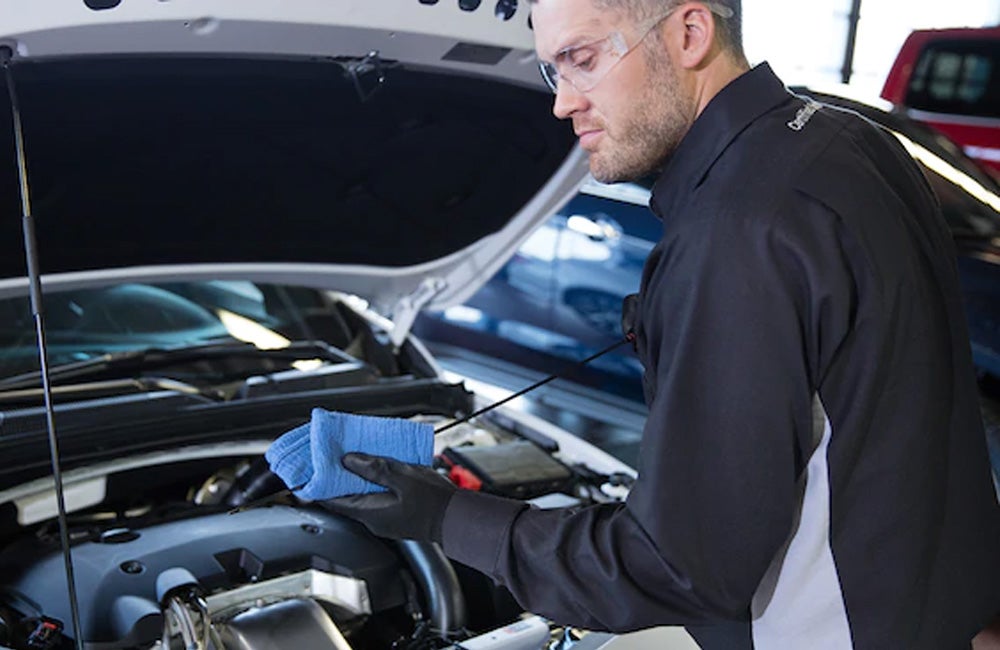 Mechanic checks under a car’s hood while holding a blue shop cloth.